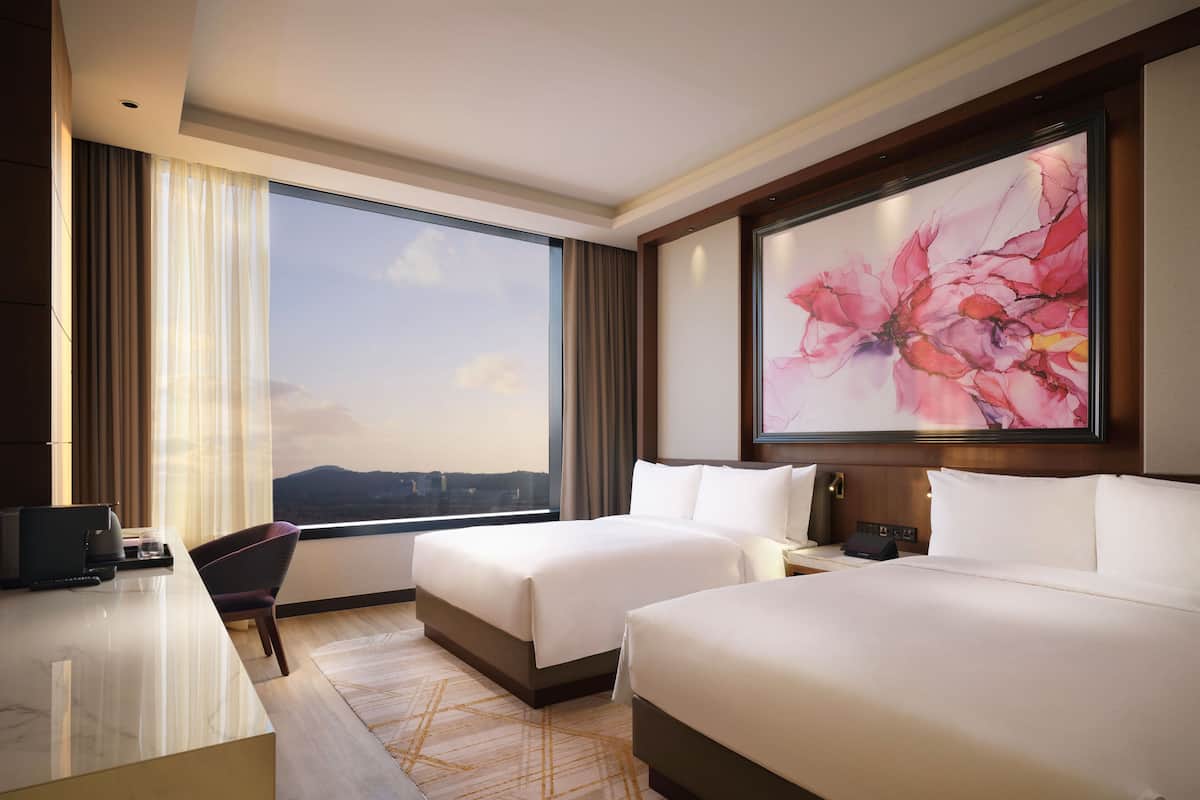 A hotel room with two beds, a desk, a chair, a large window with a city view, and a large floral artwork on the wall.