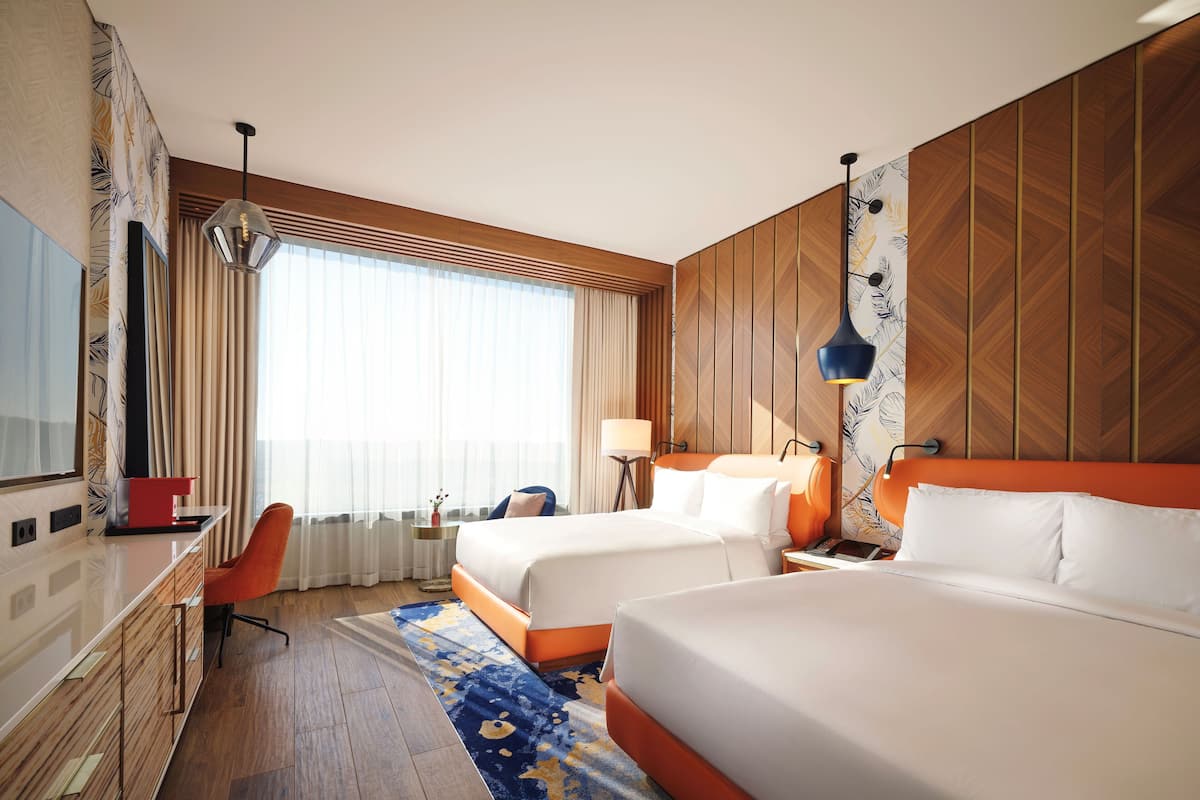 A hotel room with two beds, a desk, a chair, a TV, and a large window with curtains.