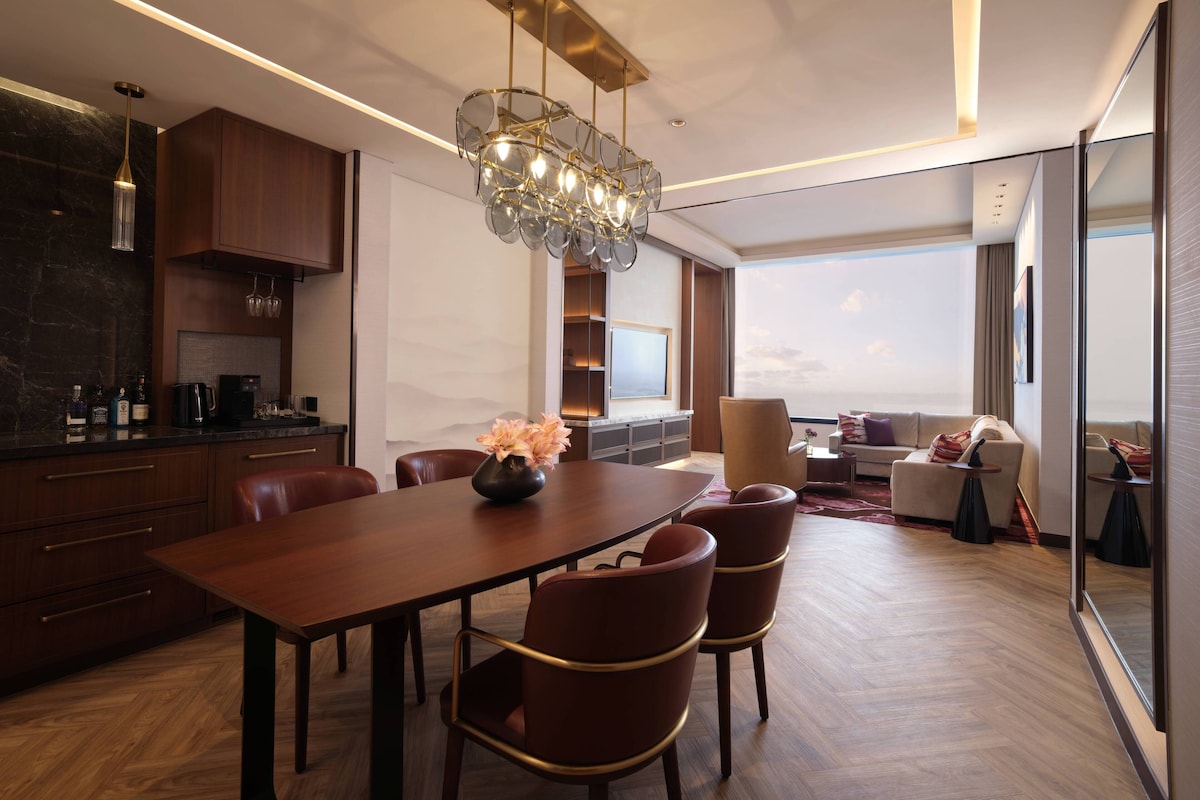 A modern hotel room with a dining area, a large window with a view, and a chandelier.