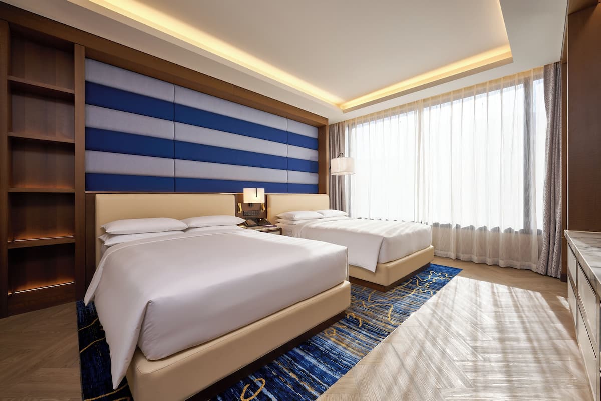 A hotel room with two beds, a large window with curtains, a blue patterned rug, and a wooden headboard.