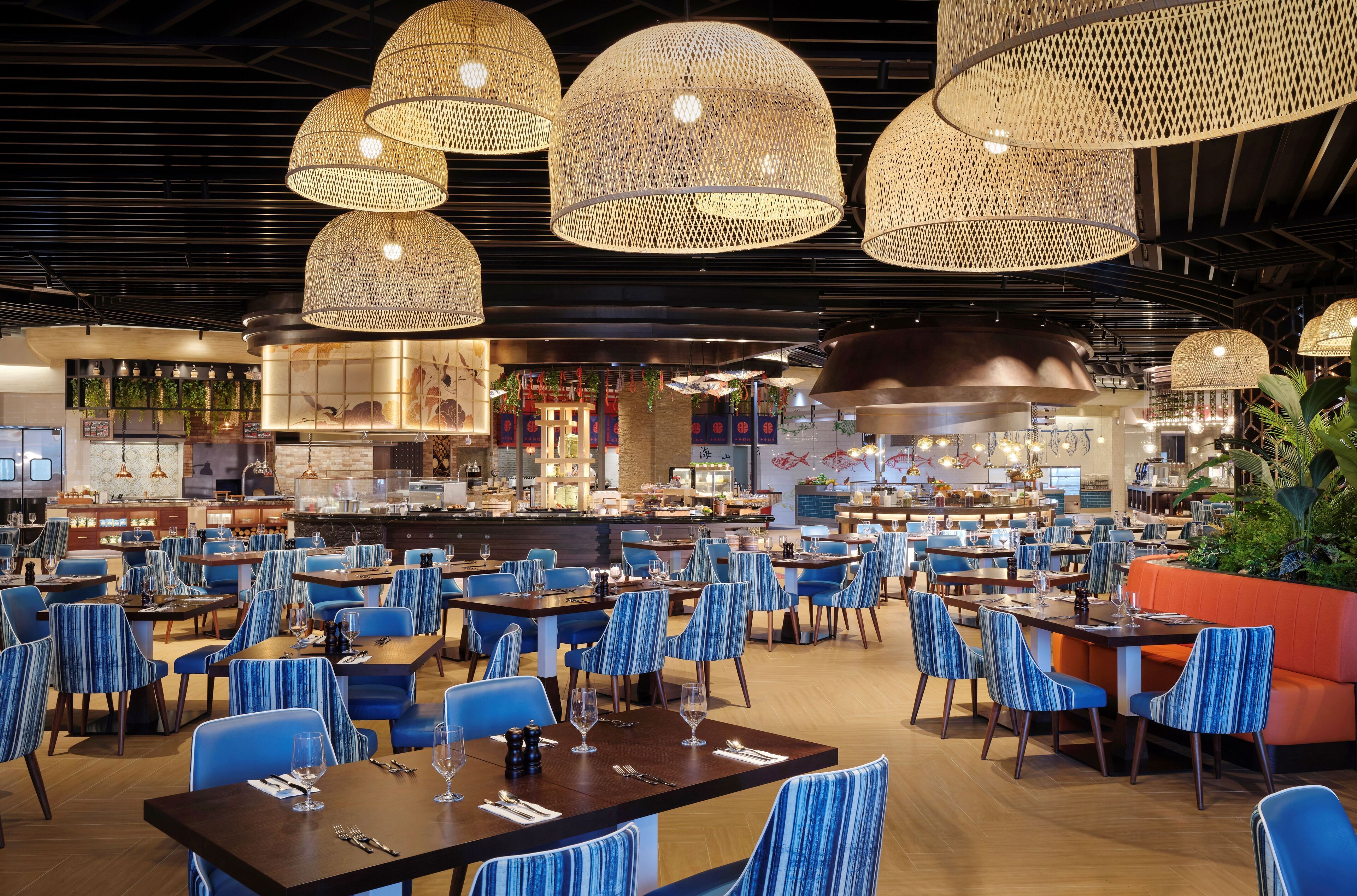 A spacious restaurant with multiple dining tables and chairs, featuring a modern interior with hanging wicker lamps and a bar area in the background.