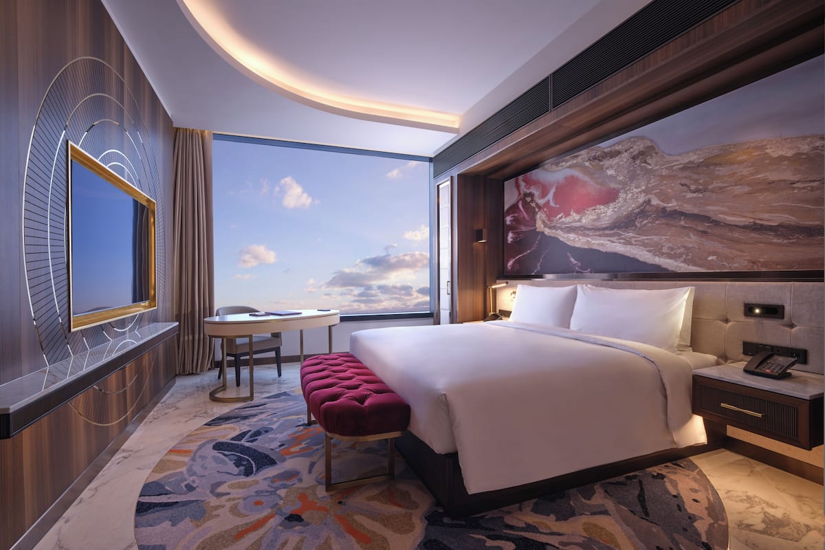 A modern hotel room with a large bed, a desk, a chair, a bench, a large window with a view, and a painting on the wall.