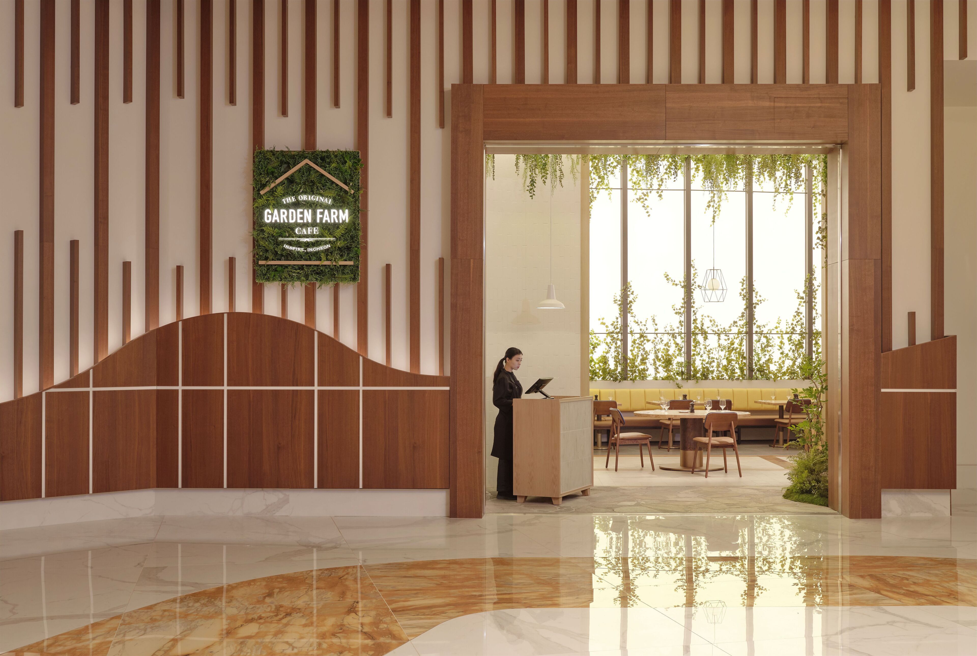A modern interior with a wooden reception desk, a sign reading "The Original Garden Farm Cafe," and a seating area with tables and chairs.