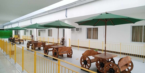 A covered outdoor seating area with wooden tables and chairs, green umbrellas, and a yellow railing. - Asia Jem Hotel-Gapan City (Gapan City)