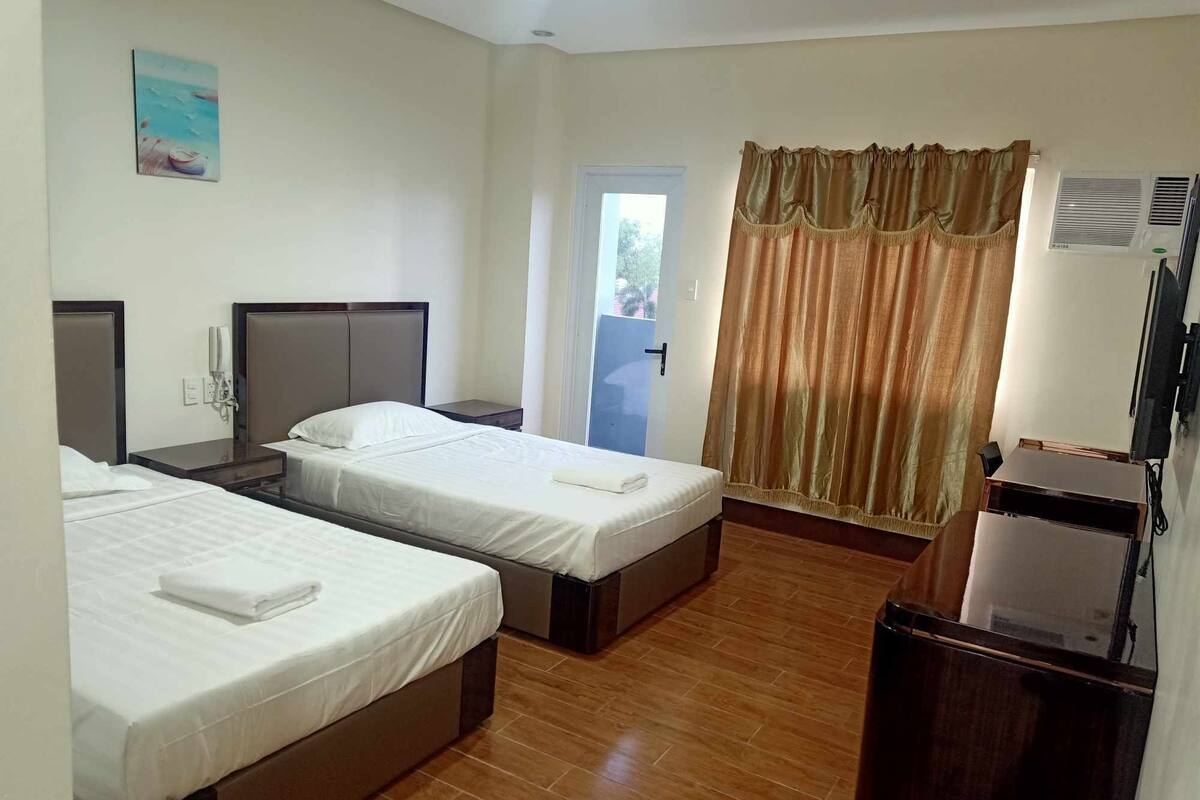 A hotel room with two beds, a wooden floor, a door with a view, and a television.