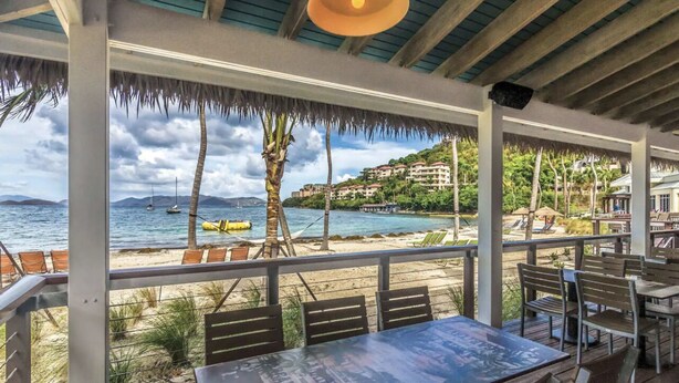 A beachside restaurant with outdoor seating, overlooking the sea and a hillside with buildings.