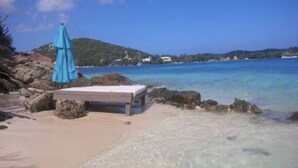 A beach with a bed and a blue umbrella, clear blue water, and a hilly landscape in the background.