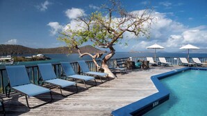 A wooden deck with lounge chairs and a pool, overlooking a body of water and distant hills.