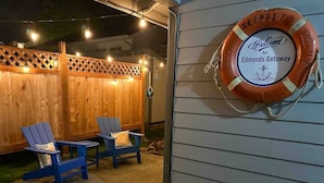A wooden deck with two Adirondack chairs and a small table, a lifebuoy with a welcome sign, and a wooden fence with string lights. - Edmonds Getaway Cottage | Walk to Train & Stadium (Edmonds)