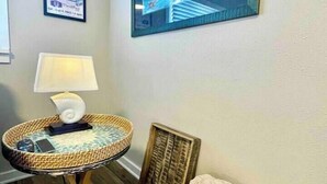A room with a wicker side table, a lamp, a framed picture, and a wicker basket with towels. - Edmonds Getaway Cottage | Walk to Train & Stadium (Edmonds)