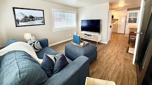 A living room with a sofa, a television, a window with blinds, and a kitchen area in the background. - Edmonds Getaway Cottage | Walk to Train & Stadium (Edmonds)
