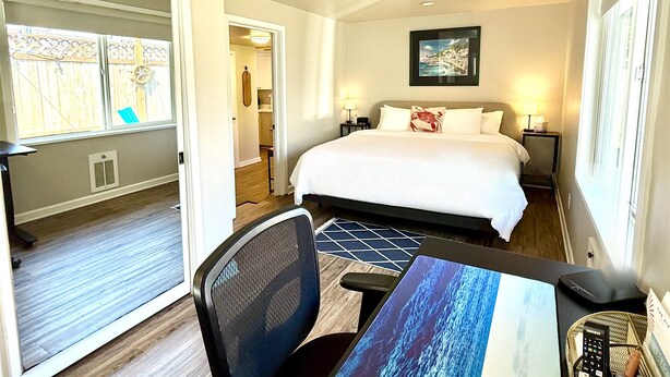A hotel room with a bed, a desk, a chair, a table, a lamp, a picture on the wall, and a window with a view of a wooden deck. - Edmonds Getaway Cottage | Walk to Train & Stadium (Edmonds)
