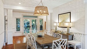 A dining room with a wooden table, white wicker chairs, a hanging light fixture, and a map on the wall. - POPULAR 4BR Silver Moss Home, Modern, Short Walk to Beach, Community Pool Access! (Kiawah Island)