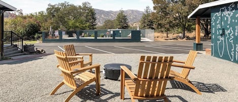 An outdoor seating area with wooden chairs and a fire pit, surrounded by a gravel ground and a fenced area with a view of mountains.