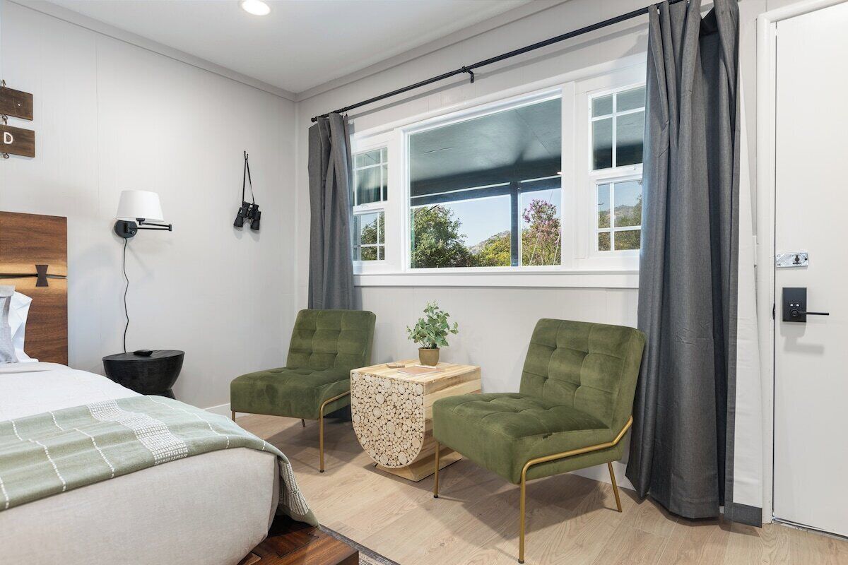 Une chambre d’hôtel avec un lit, deux fauteuils verts, une petite table avec une plante, une fenêtre avec des rideaux et une lampe fixée au mur.