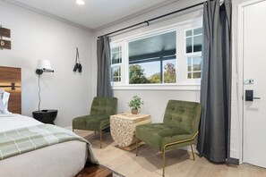 A hotel room with a bed, two green armchairs, a small table with a plant, a window with curtains, and a wall-mounted lamp. - The Atwell at the Sequoia Motel (Three Rivers)