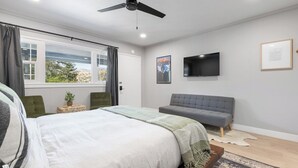 Un dormitorio con una cama, un sofá gris, un televisor en la pared y una ventana con cortinas.