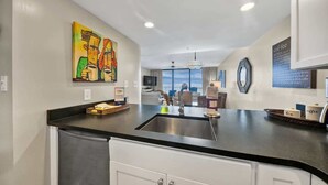 A modern kitchen with a black countertop, stainless steel sink, and white cabinets. There is a painting on the wall and a coffee sign. - Newly Updated 2BR Beachfront Condo With Balcony and Direct Beach Access (Santa Rosa Beach)