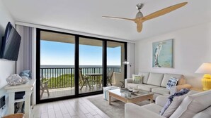 A spacious living room with a large sliding glass door leading to a balcony, a ceiling fan, a flat-screen TV, a sofa set, and a painting on the wall. - One Seagrove Place 0208 (Santa Rosa Beach)