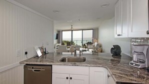 A modern kitchen with a granite countertop, stainless steel sink, and white cabinetry. There is a coffee maker on the counter and a dishwasher below. - One Seagrove Place 0208 (Santa Rosa Beach)