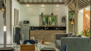 A dining area with a table set for two, a kitchen with a refrigerator and microwave, and a view of the outdoors. - Mundaka Villa by Betterplace (Canggu)