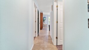 A hallway with white walls and beige tiled flooring, leading to a room with a television and a chair.