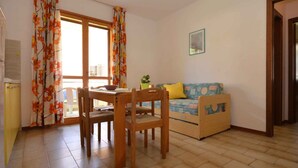 A cozy living room with a wooden dining table, chairs, a sofa with a cushion, and a window with curtains. - Holiday apartment with balcony and air conditioning (Lignano)