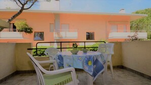 A patio with a table set for two, surrounded by white chairs, overlooking a building with orange walls and balconies. - Holiday apartment with balcony and air conditioning (Lignano)
