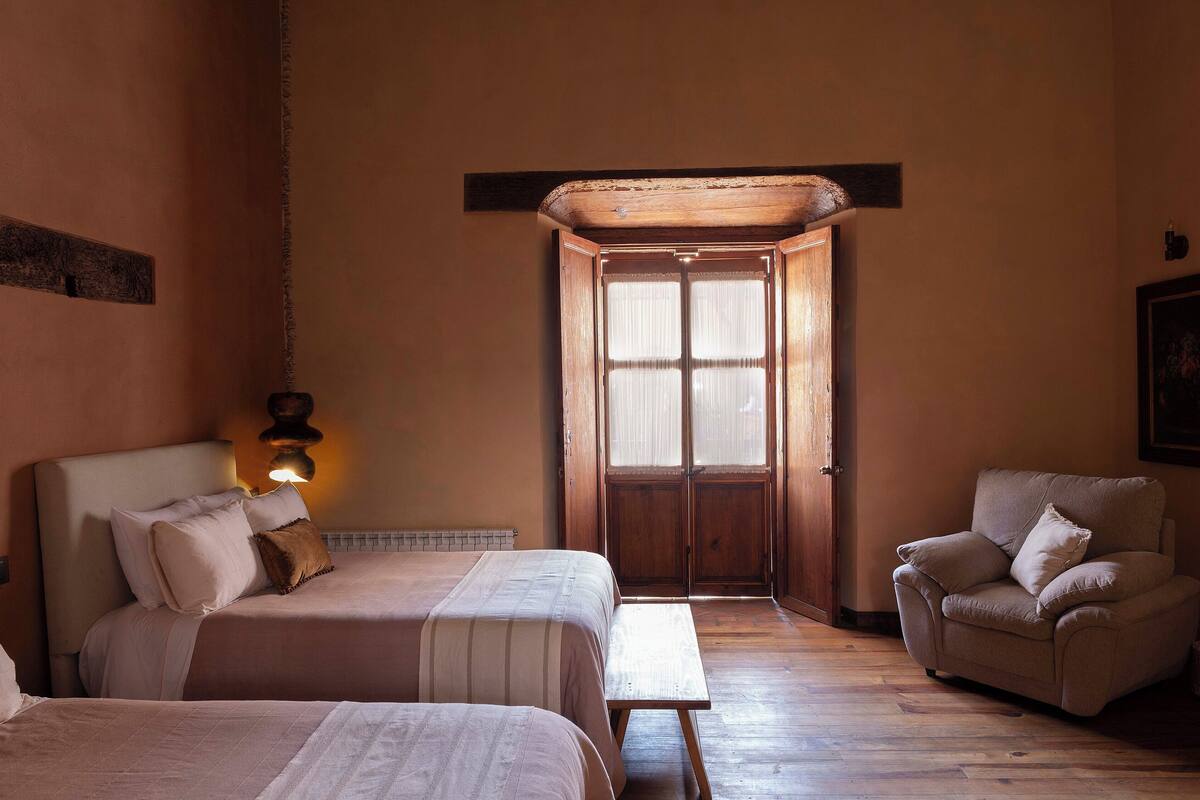 A room with two beds, a wooden bench, a chair, and a door leading to another room.