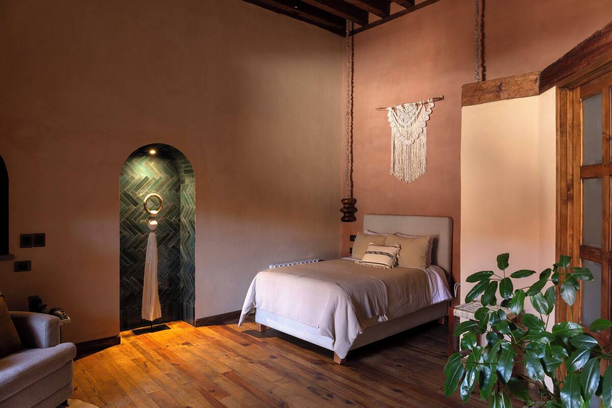 A bedroom with a bed, wooden flooring, a hanging macrame piece, and a plant.
