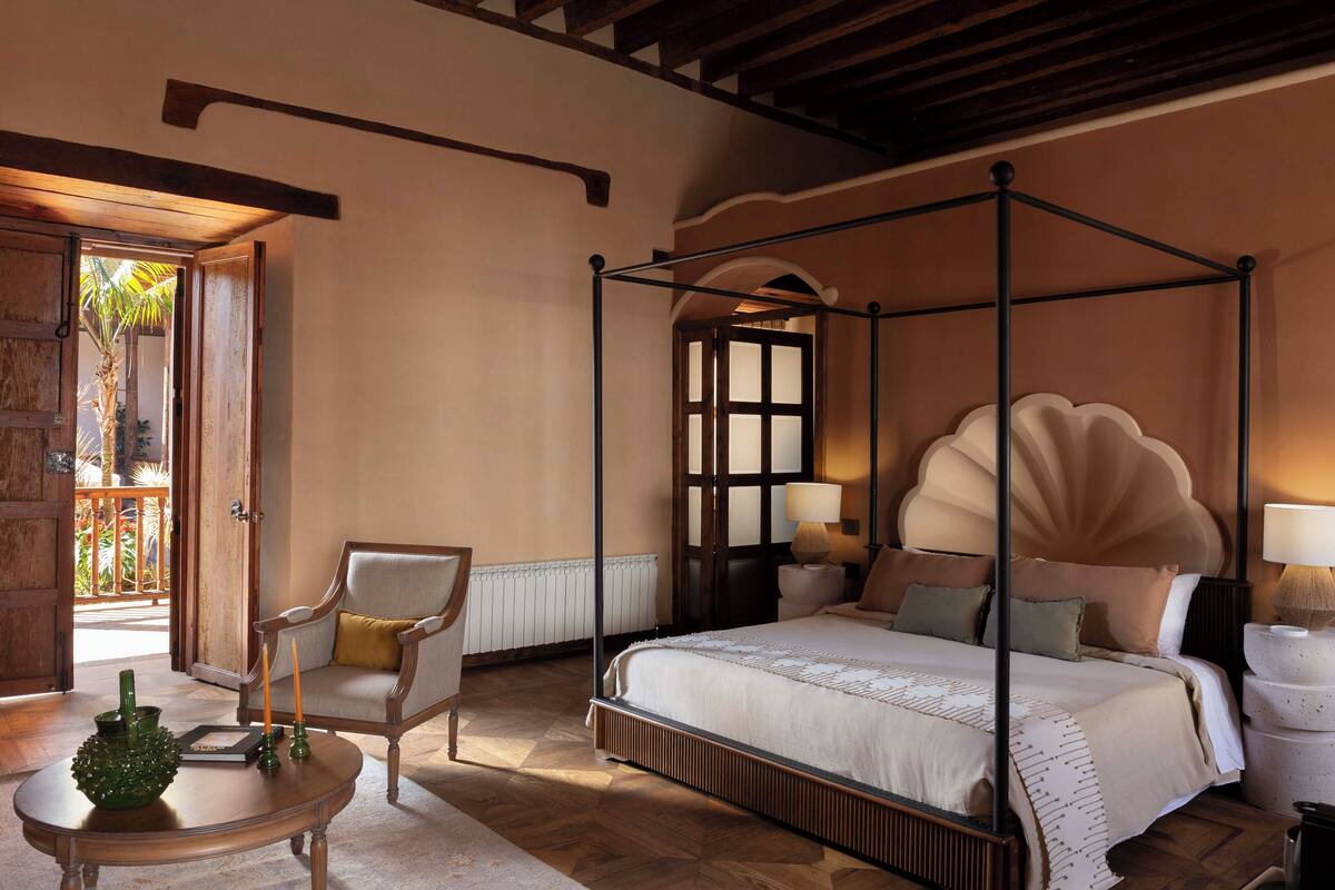A four-poster bed with a canopy, a sitting chair, a small table with candles, and a view of the outdoors through a door.