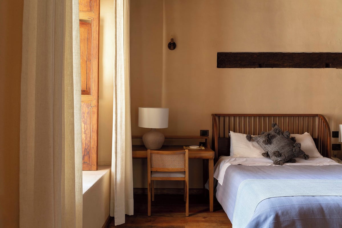 A bedroom with a wooden desk, a chair, a bed with white linens, and a window with curtains.