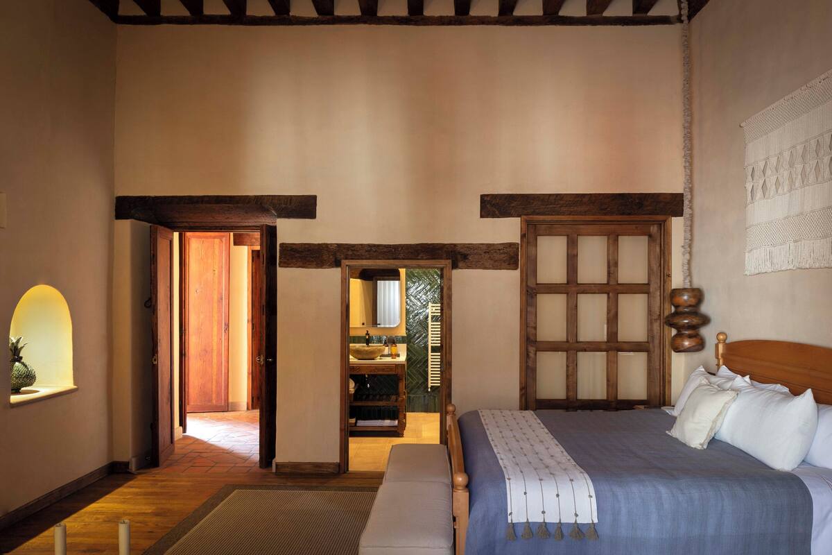 A bedroom with a wooden bed, a bench, a small window with a plant, and a doorway leading to a bathroom with a sink and mirror.