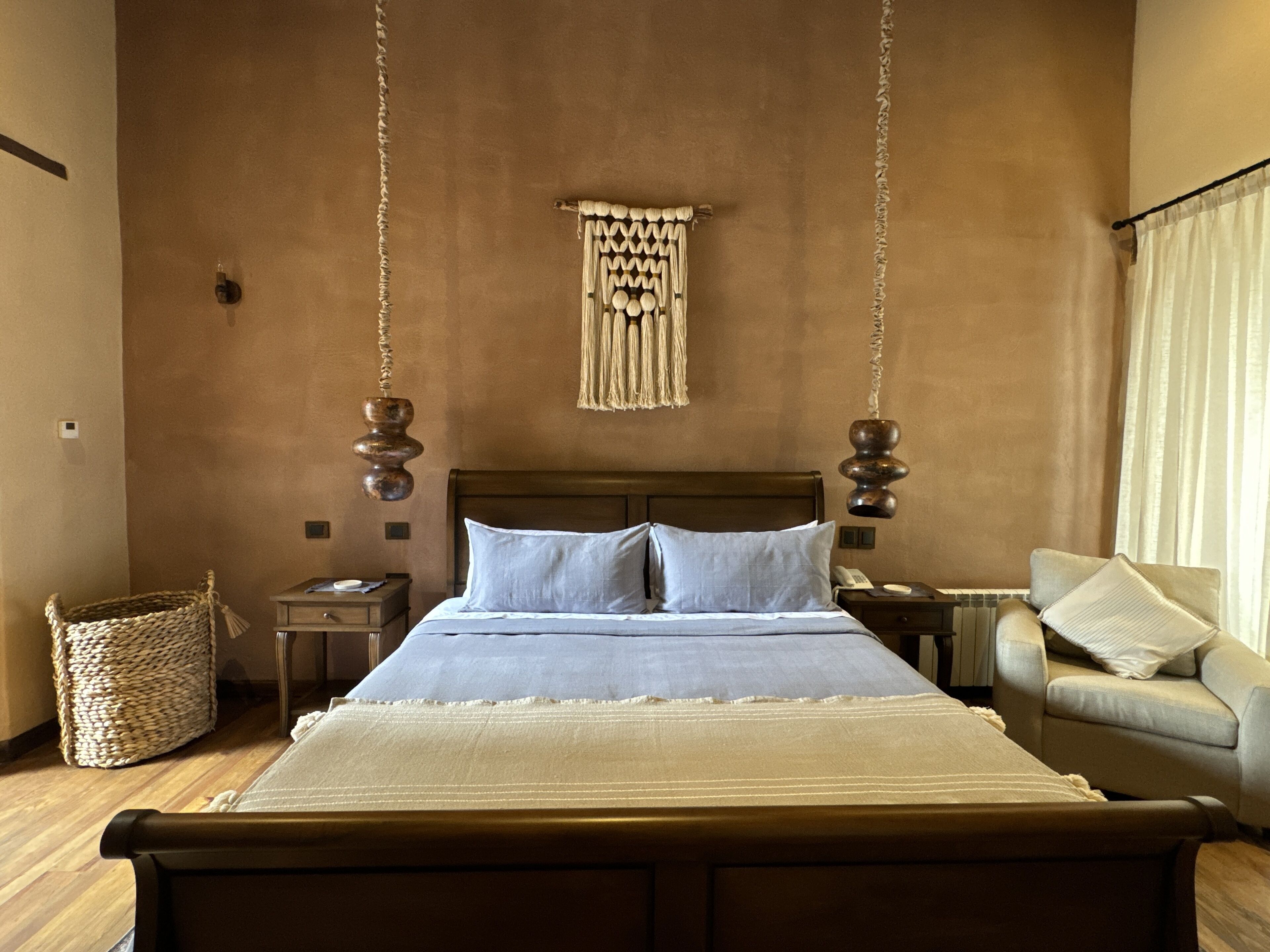 Un dormitorio con una cama grande, cabecera de madera, decoración de macramé colgante, una cesta tejida y un sofá.
