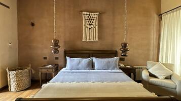 Un dormitorio con una cama grande, cabecera de madera, decoración de macramé colgante, una cesta tejida y un sofá.