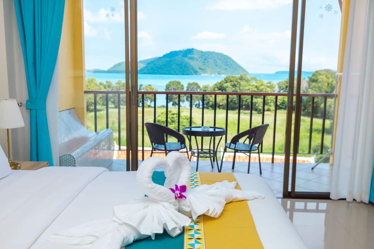 Une chambre d’hôtel avec un lit, un balcon donnant sur une montagne et l’eau, et deux chaises.