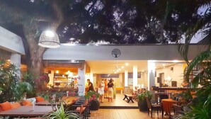 An outdoor seating area with wooden tables and chairs, surrounded by potted plants and a large tree. - Kana Hotel Phuket (Rawai)