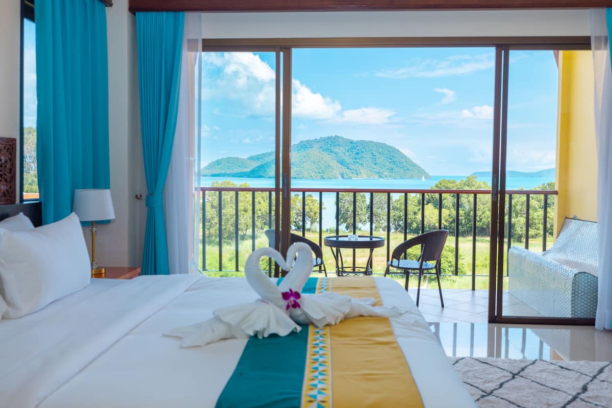 Une chambre d’hôtel avec un grand lit, un balcon offrant une vue sur la montagne et la mer, ainsi qu’une table avec des chaises à l’extérieur.