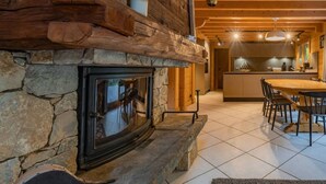 A stone fireplace with a wooden mantel, a tiled floor, and a dining area with a table and chairs. - Chalet Glaciers | Fireplace | View | Comfy | Quiet (Les Houches)