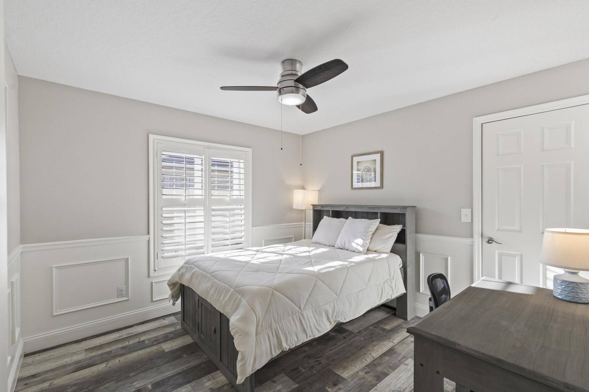 A bedroom with a bed, a ceiling fan, a window with blinds, a bedside lamp, and a door.