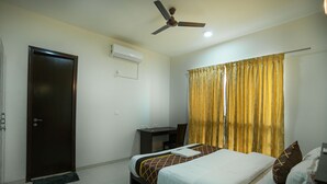 A hotel room with a bed, a desk, a ceiling fan, and a window with curtains. - Urban Stay Service Apartment (Chengalpattu)