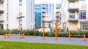 A modern urban street with a pedestrian walkway, a metal bench, and a building with balconies and large windows. - The Hunt Cherrywood (Laughanstown)
