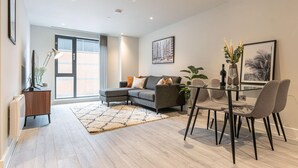 A modern living room with a sectional sofa, a glass dining table with chairs, a large window, and a radiator. - St Martin's Place - Birmingham City Centre (Birmingham)
