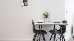 A modern dining area with a white table, two black chairs, a small potted plant, and a wall-mounted phone. - Sevenstays Residences Solihull - NEC / Airport (Solihull)