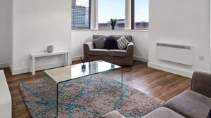 Uma sala de estar moderna com uma mesa de centro de vidro, um sofá cinzento e vista para a cidade através de amplas janelas.
