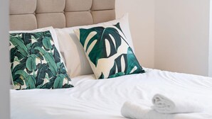 Cama bem arrumada com lençóis brancos, duas almofadas com estampas de folhas tropicais e cabeceira estofada bege.