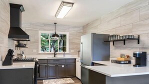 A modern kitchen with dark cabinets, a stainless steel refrigerator, a coffee maker, and a window with a view of greenery. - Brookside- One of a Kind (Tulsa)
