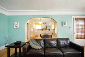 A living room with a black leather sofa, a small table, and a vase with flowers. There is a doorway leading to a kitchen and dining area.
