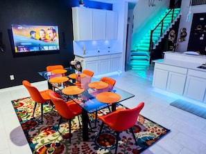 A modern kitchen with white cabinets, a glass-top dining table with orange chairs, and a colorful rug.