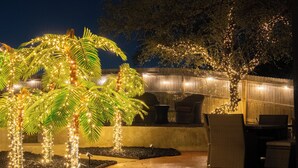 Un patio al aire libre con palmeras adornadas con luces colgantes, una valla de madera y mobiliario de mimbre.